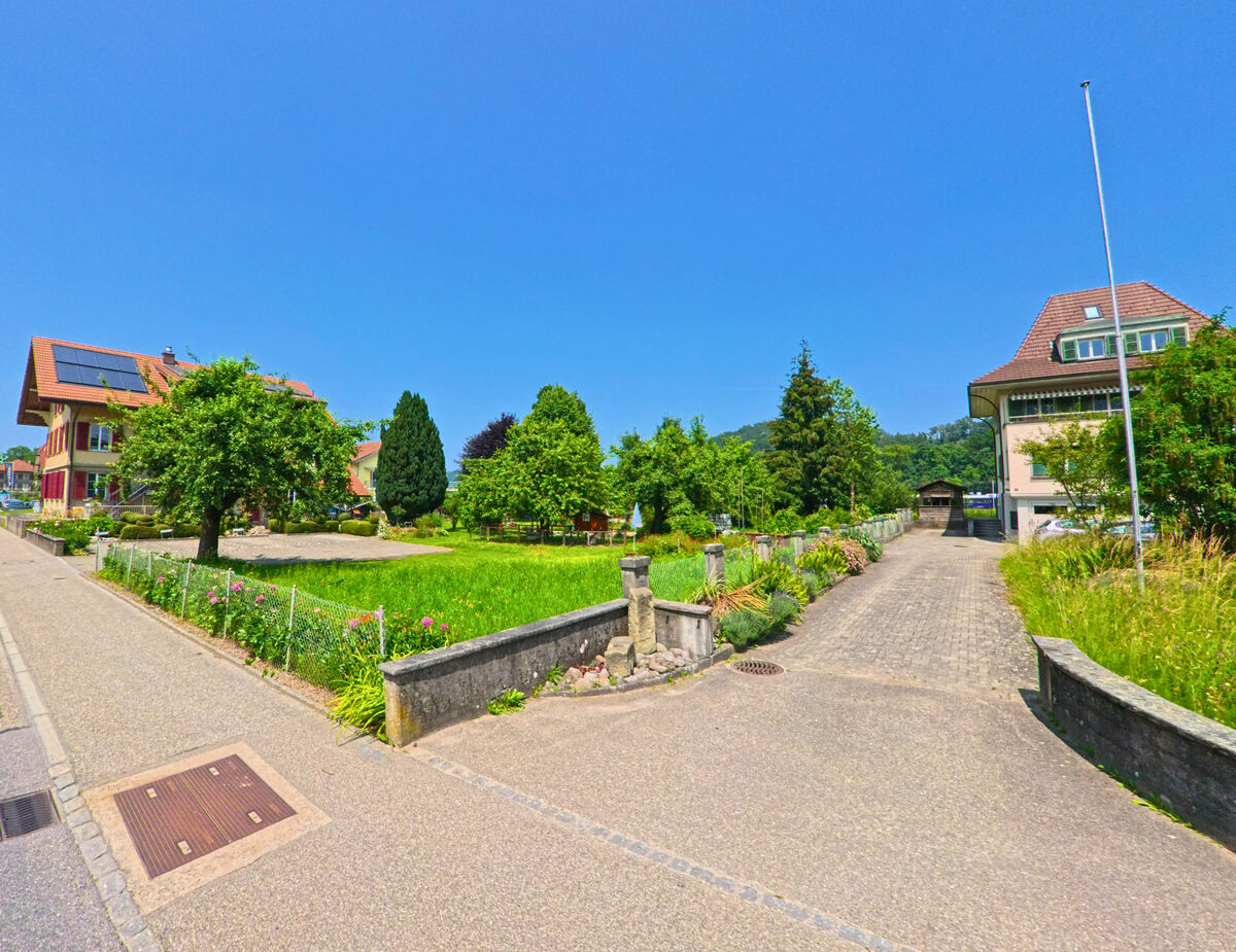 In Oberburg (PLZ 3414, Kanton Bern) führt ein gepflasterter Weg zwischen gepflegten Vorgärten und zwei Wohnhäusern mit roten Ziegeldächern durch ein ruhiges Quartier.