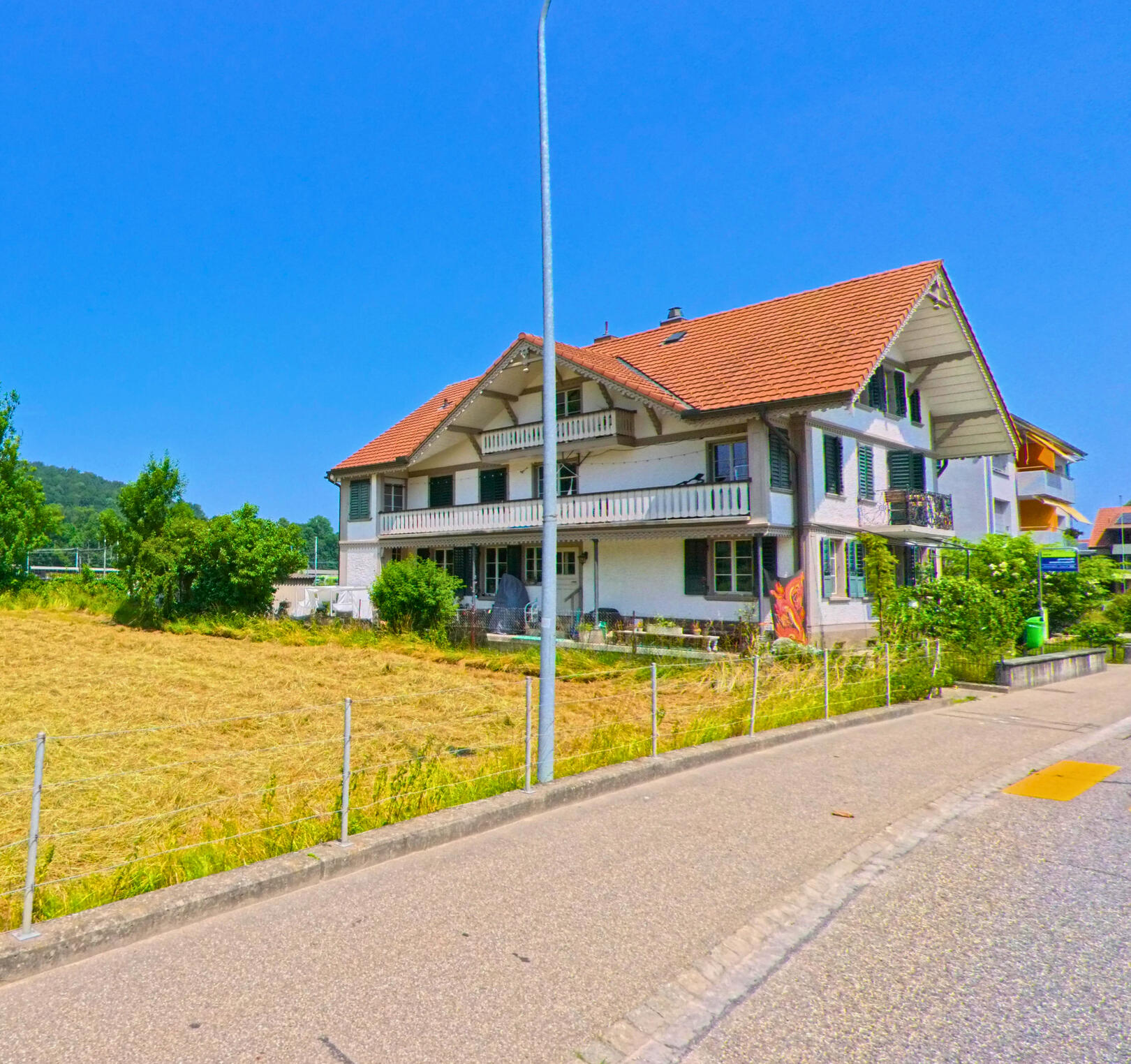 In Oberburg (3414, Kanton Bern) steht ein weiss verputztes Mehrfamilienhaus mit roten Ziegeldächern und Balkonen direkt an einer ruhigen Quartierstrasse.