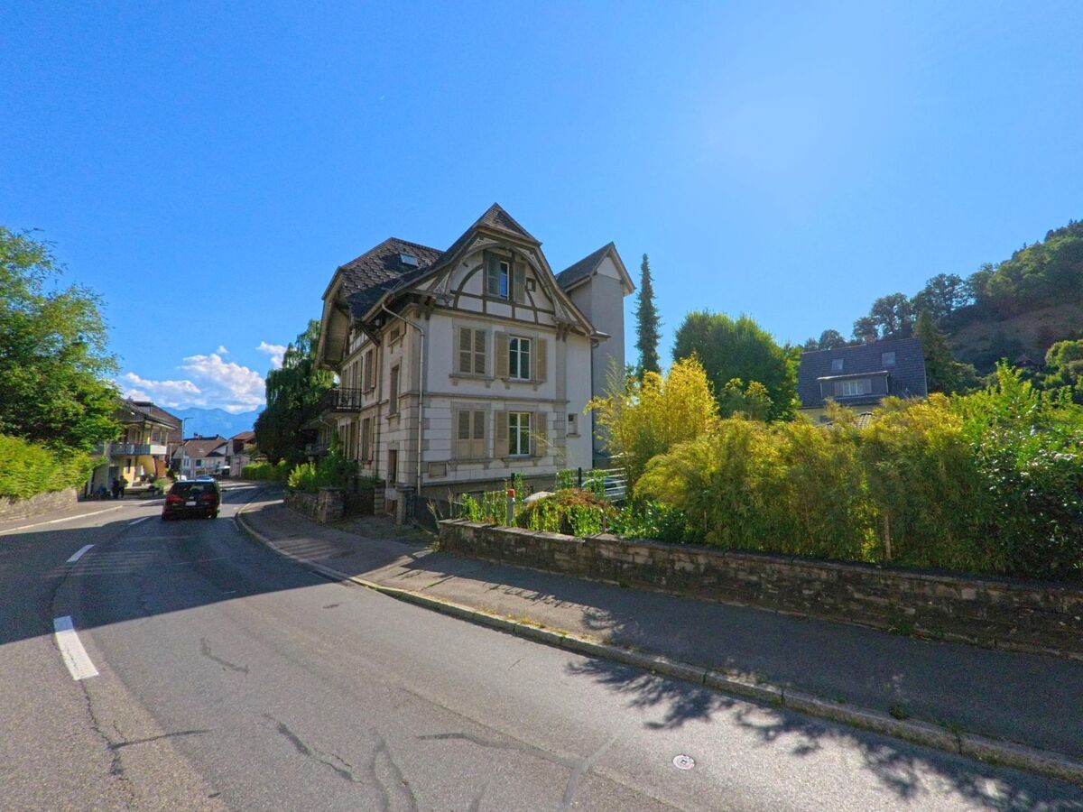 In Oberdiessbach (3672, Kanton Bern) verläuft eine kurvige Strasse an einem eleganten Altbau mit Giebeldach vorbei, der von dichtem Grün und einem Naturgarten umgeben ist.