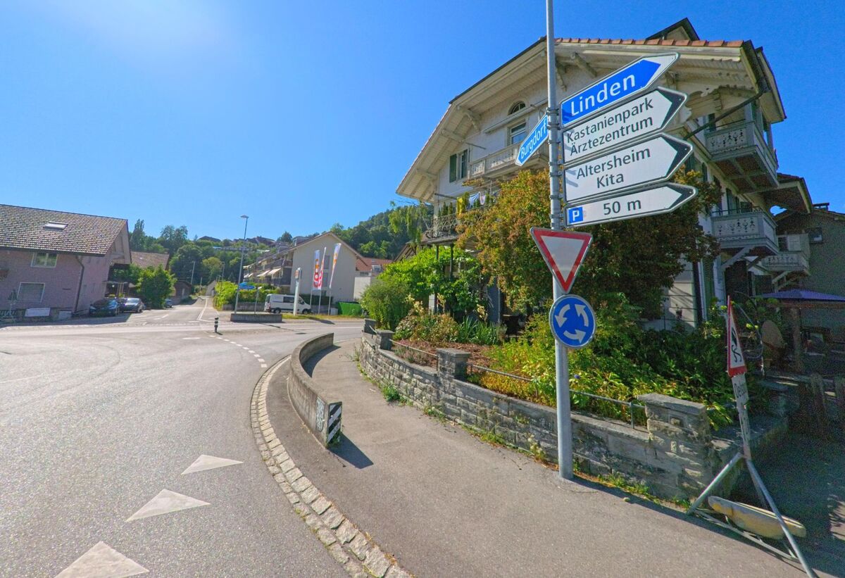 In Oberdiessbach (3672, Kanton Bern) steht an einem kleinen Kreisel ein Wegweiser vor einem Haus mit Balkon und Garten, der auf Einrichtungen wie Arztzentrum und Altersheim hinweist.