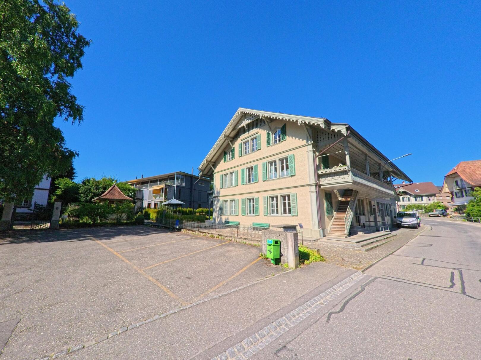 In Oberdiessbach (3672, Kanton Bern) steht ein grosses Haus mit grünen Fensterläden und verziertem Holzbalkon an einer ruhigen Seitenstrasse neben einem leeren Parkplatz.