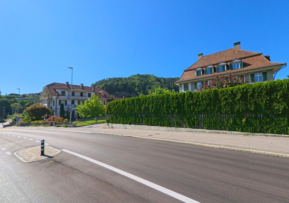 In Oberdiessbach (3672, Kanton Bern) säumen zwei grosse Häuser mit steilen Dächern eine frisch asphaltierte Strasse, getrennt durch einen dichten Sichtschutz aus hohen Hecken.