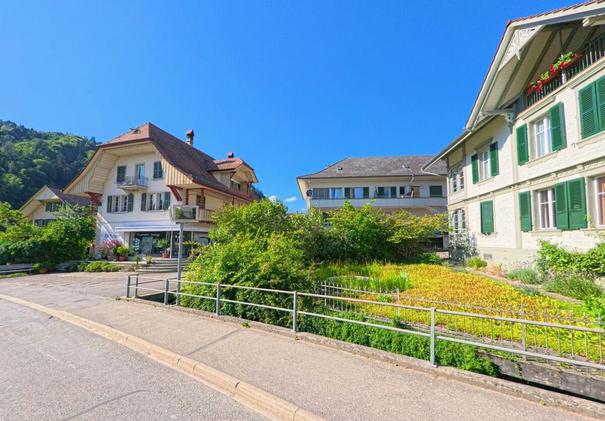In Oberdiessbach (3672, Kanton Bern) reihen sich an einer leicht gebogenen Strasse Wohnhäuser mit grünen Fensterläden und ein kleines Geschäft mit bepflanztem Vorgarten.