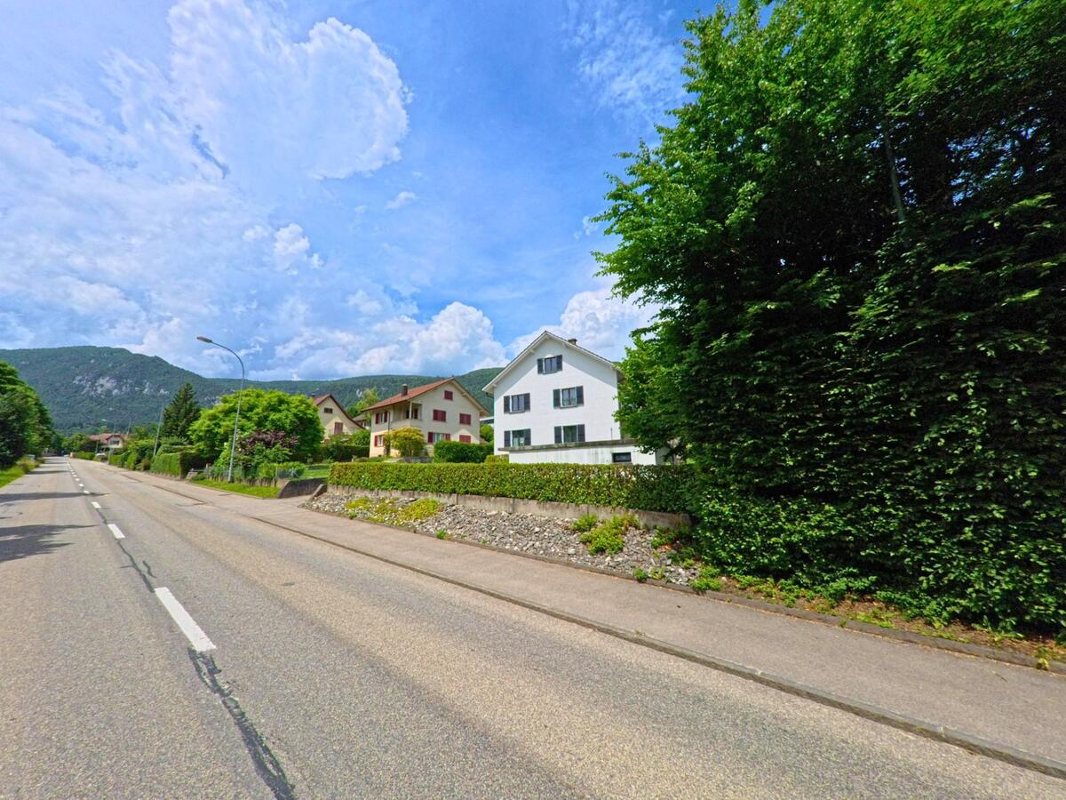 In Oberdorf (4515, Kanton Solothurn) säumen Einfamilienhäuser mit gepflegten Gärten eine breite Ausfallstrasse mit Blick auf die bewaldeten Jurahänge.