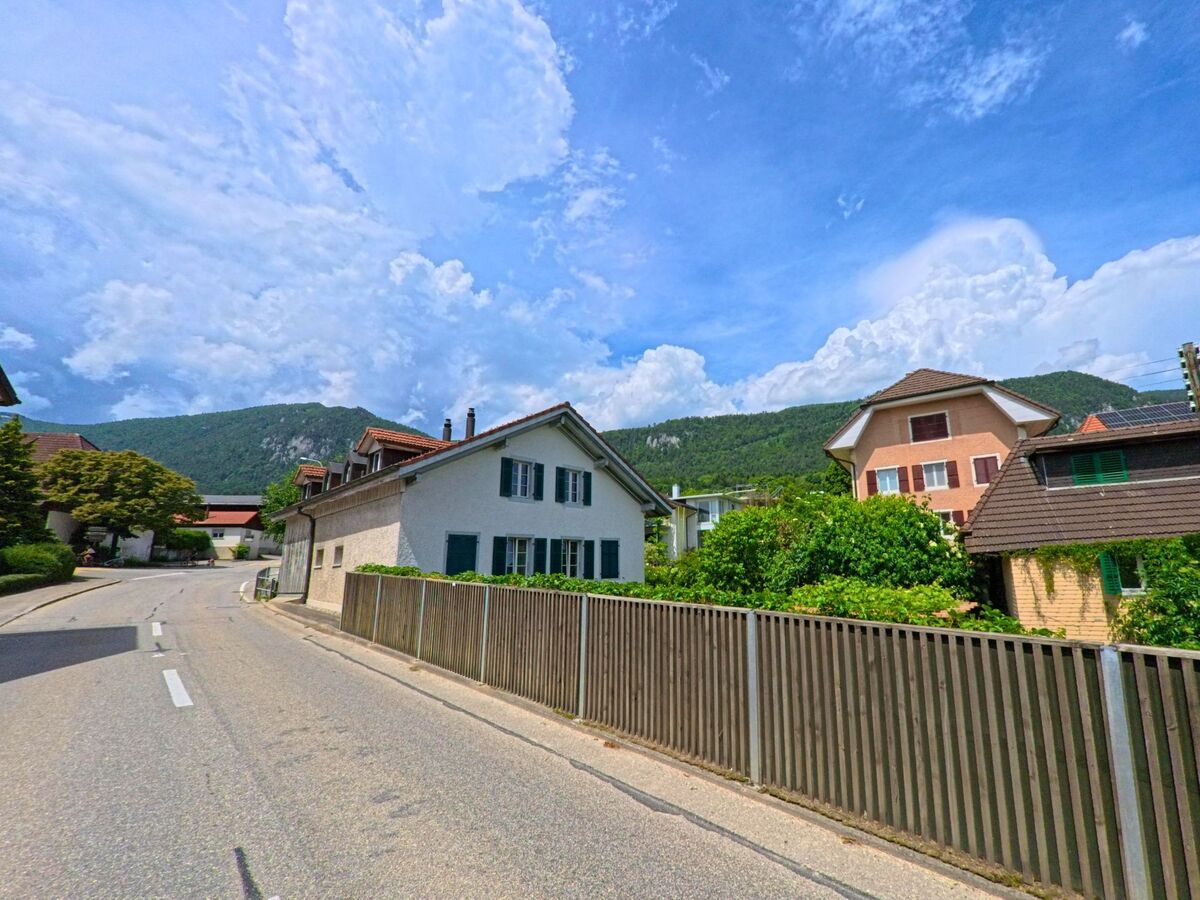 In Oberdorf (4515, Kanton Solothurn) fuehrt eine Dorfstrasse an traditionellen Wohnhaeusern vorbei, eingerahmt von einem Holzzaun und gruenen Huegeln.