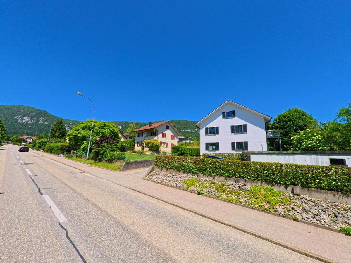 Entlang einer leicht ansteigenden Strasse in Oberdorf (4515, Kanton Solothurn) stehen gepflegte Wohnhäuser mit Hecken und Gärten vor bewaldeter Kulisse.