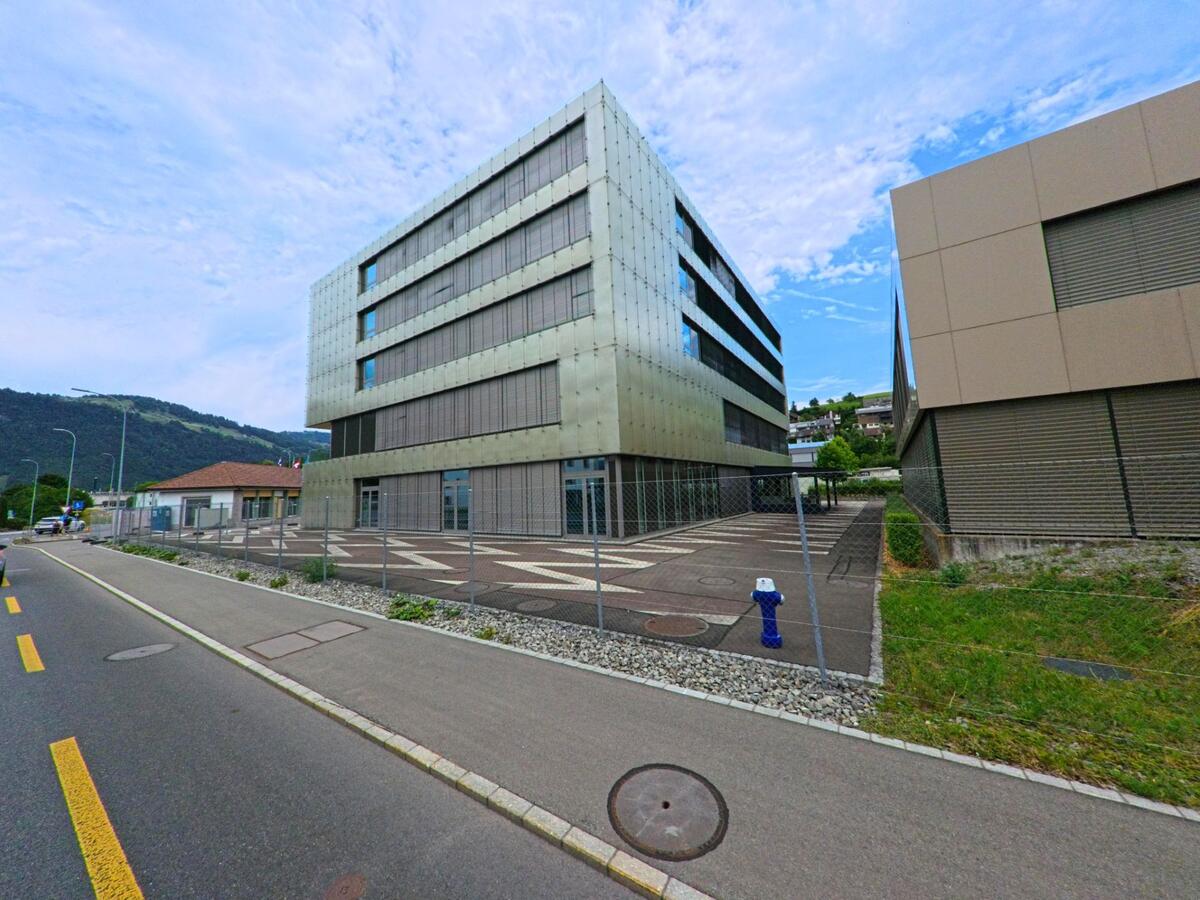 In Oberdorf (6370, Kanton Nidwalden) steht ein modernes, mehrgeschossiges Gebaeude mit metallisch wirkender Fassade neben einer verkehrsarmen Strasse und angrenzendem Gehweg.