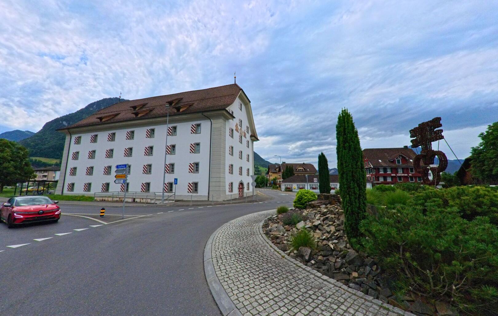 In Oberdorf (6370, Kanton Nidwalden) steht ein markantes weisses Gebaeude mit rot-weiss gestrichenen Fensterlaeden an einer Strassengabelung, daneben eine gestaltete Verkehrsinsel mit Bepflanzung und Skulptur.