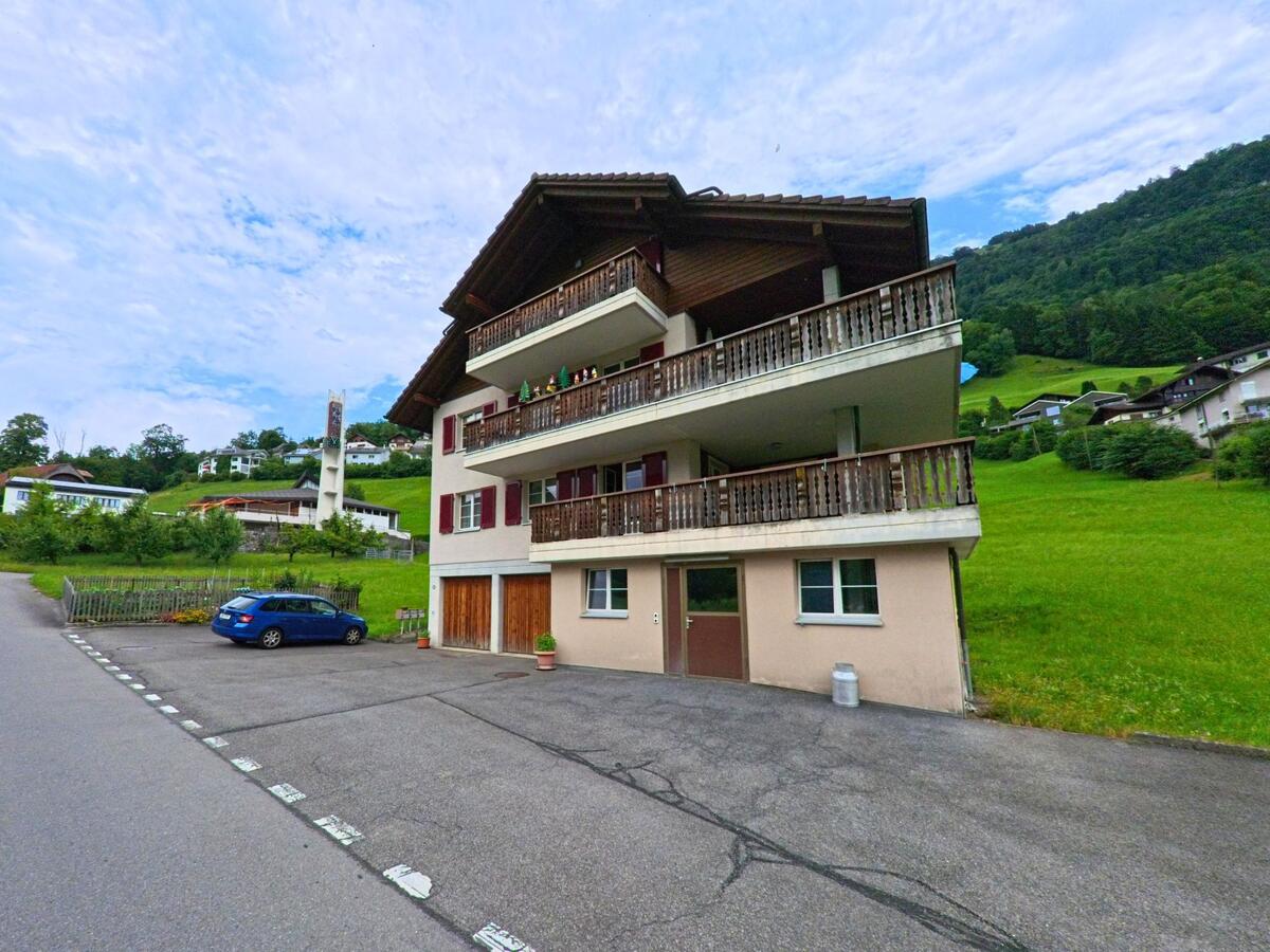 In Oberdorf (6382, Kanton Nidwalden) steht ein Wohnhaus mit Holzverkleidung und breiten Balkonen am Hang, im Hintergrund ragt ein markanter Kirchturm aus der Umgebung hervor.