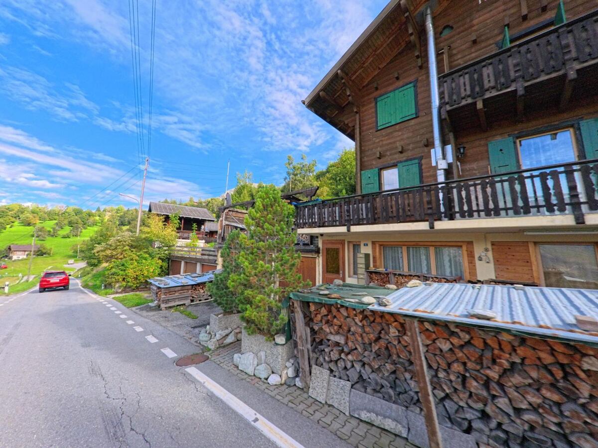 Entlang einer schmalen Strasse in Oberems (3948, Kanton Wallis) stehen traditionelle Holzhäuser mit grünen Fensterläden und gestapeltem Brennholz.