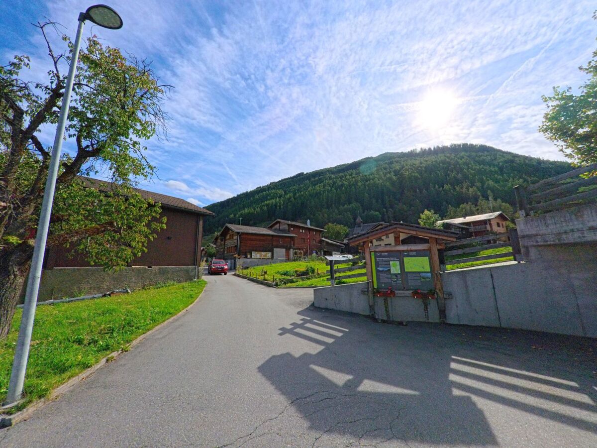 In Oberems (3948, Kanton Wallis) führt eine schmale Strasse mit Informationstafel und Holzhäusern bergauf Richtung eines bewaldeten Hangs.