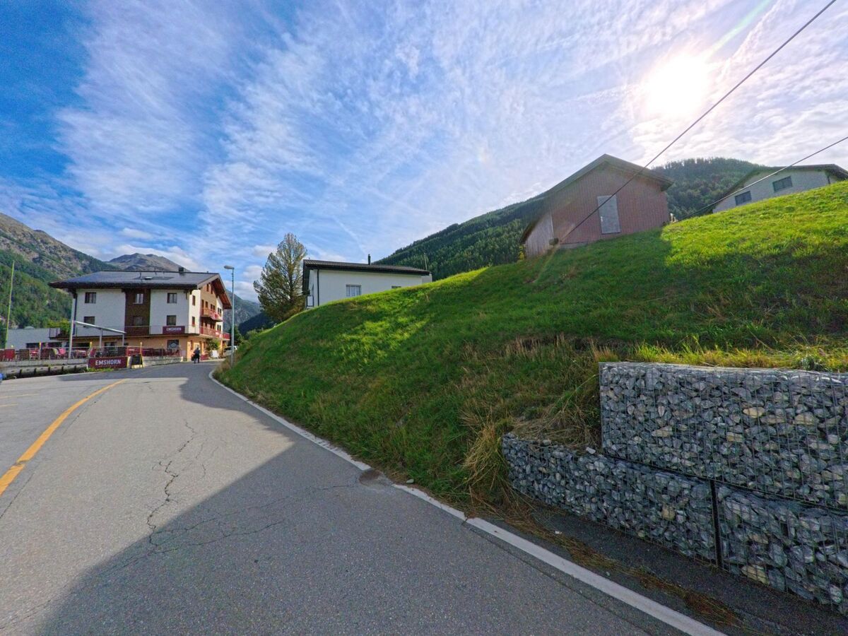 Eine leicht ansteigende Strasse in Oberems (3948, Kanton Wallis) führt vorbei an Wiesen und dem Hotel Emshorn im Sonnenschein.