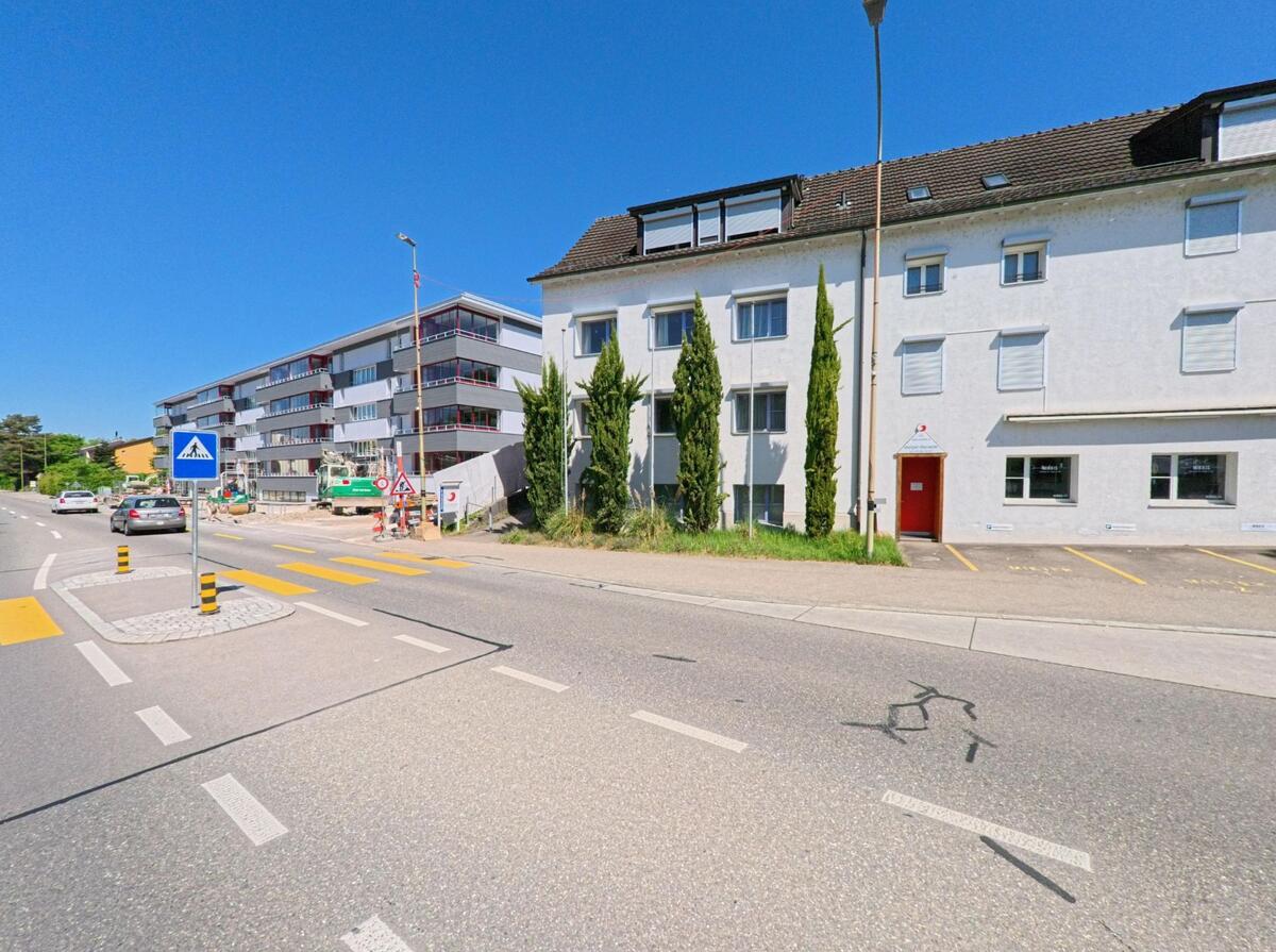  In Oberengstringen (8102, Kanton Zürich) führt ein Zebrastreifen mit Mittelinsel an einem weissen Wohnhaus und einem modernen Neubau mit Balkonen vorbei.