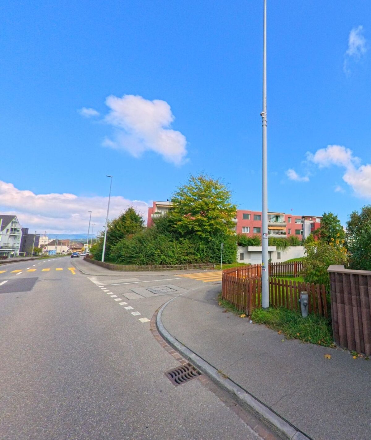 Eine Strasse in Oberentfelden (5036, Kanton Aargau) fuehrt an modernen Mehrfamilienhaeusern und dichtem Gruen vorbei, waehrend ein Fussgaengerstreifen den Weg kreuzt.