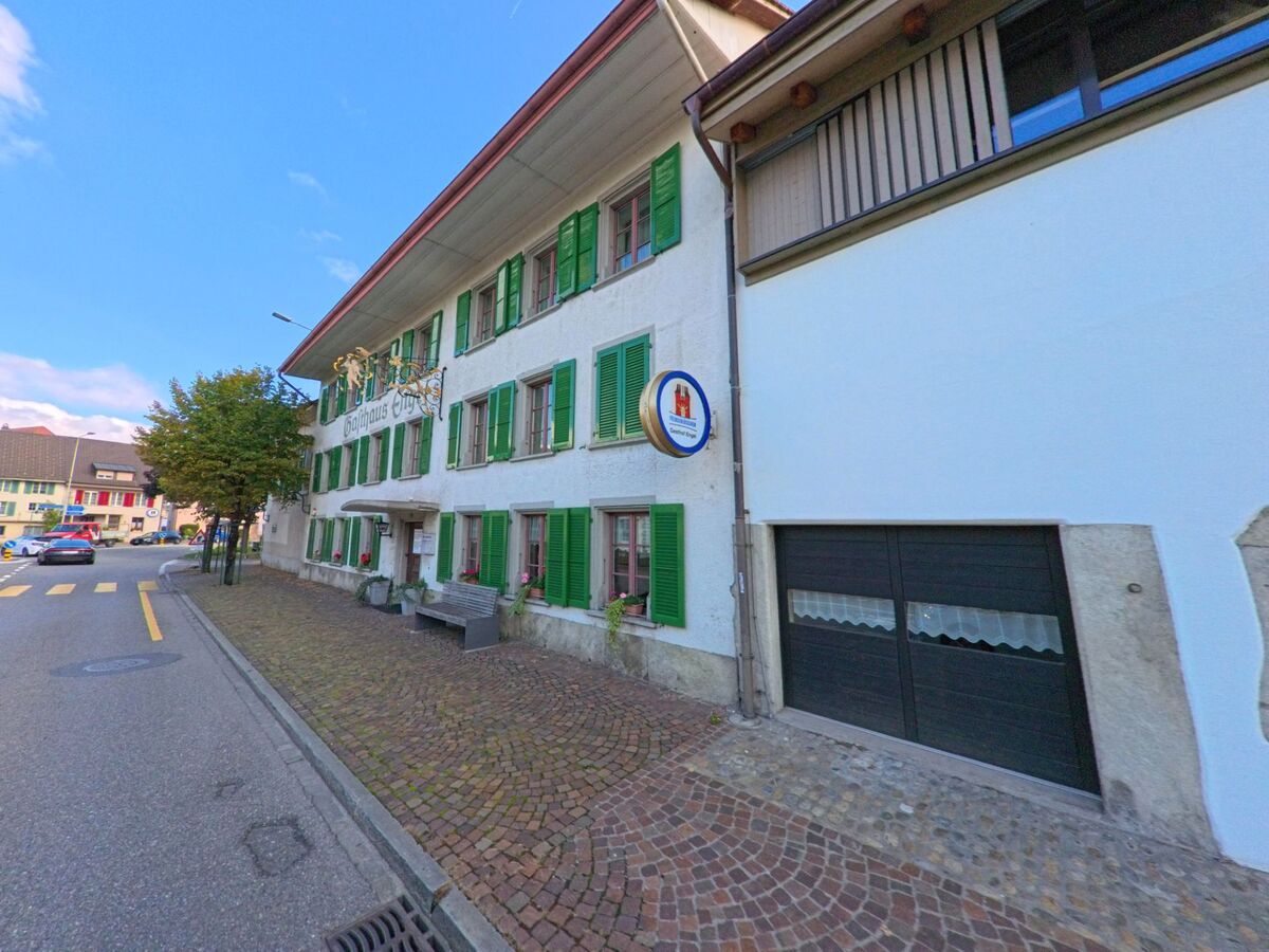 Das Gasthaus Engel in Oberentfelden (5036, Kanton Aargau) zeigt eine traditionelle Fassade mit gruenen Fensterlaeden und einem goldenen Wirtshausschild an der Strasse.