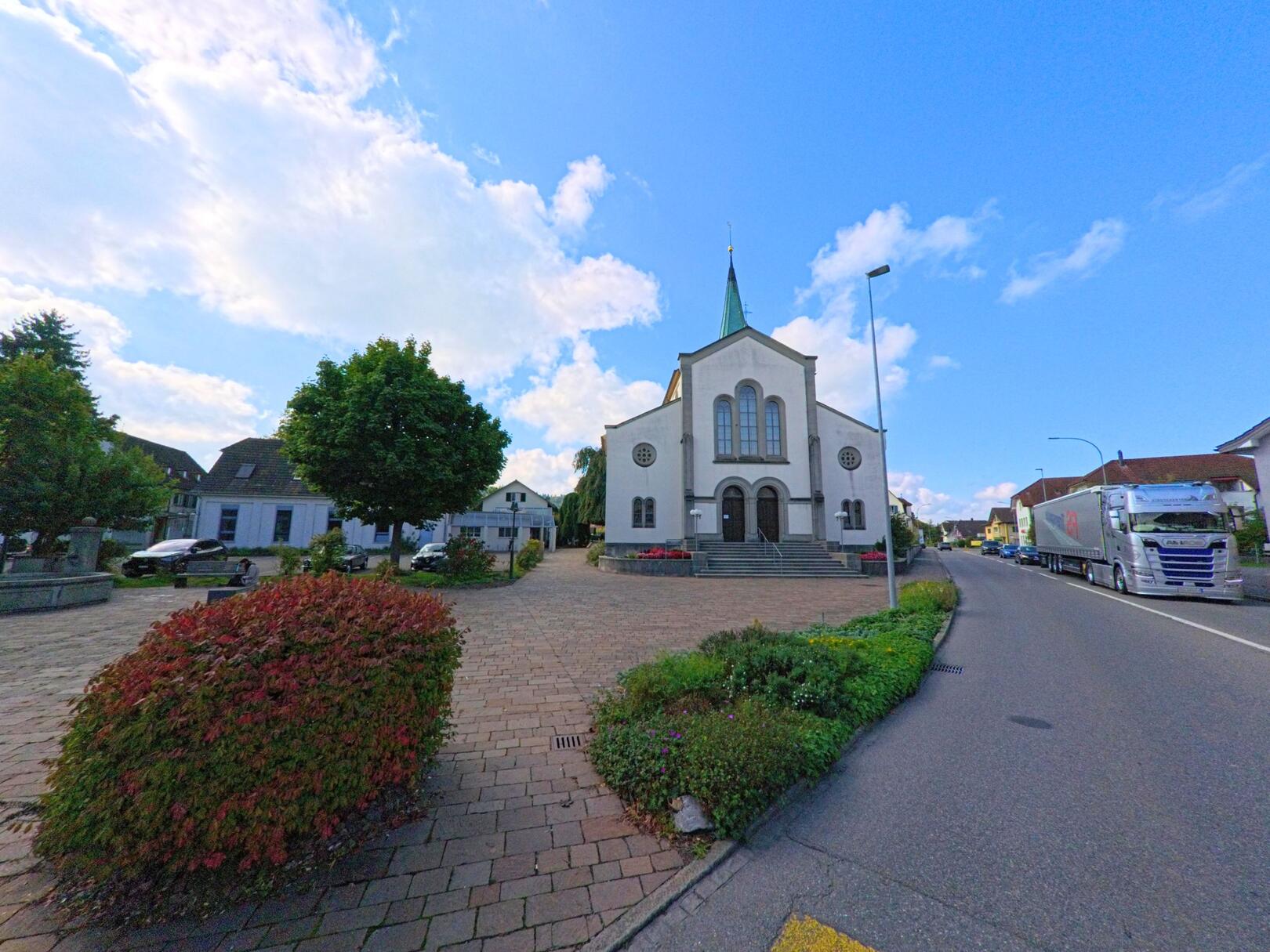 Vor der Kirche in Oberentfelden (5036, Kanton Aargau) breitet sich ein gepflasterter Platz mit Baeumen und gepflegten Pflanzen aus, waehrend die Hauptstrasse rechts vorbeifuehrt.