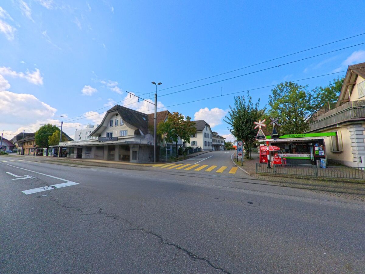 An der Hauptstrasse in Oberentfelden (5036, Kanton Aargau) befinden sich eine Tramhaltestelle, ein kleiner Kiosk und traditionelle Wohnhaeuser mit Zebrastreifen im Vordergrund.