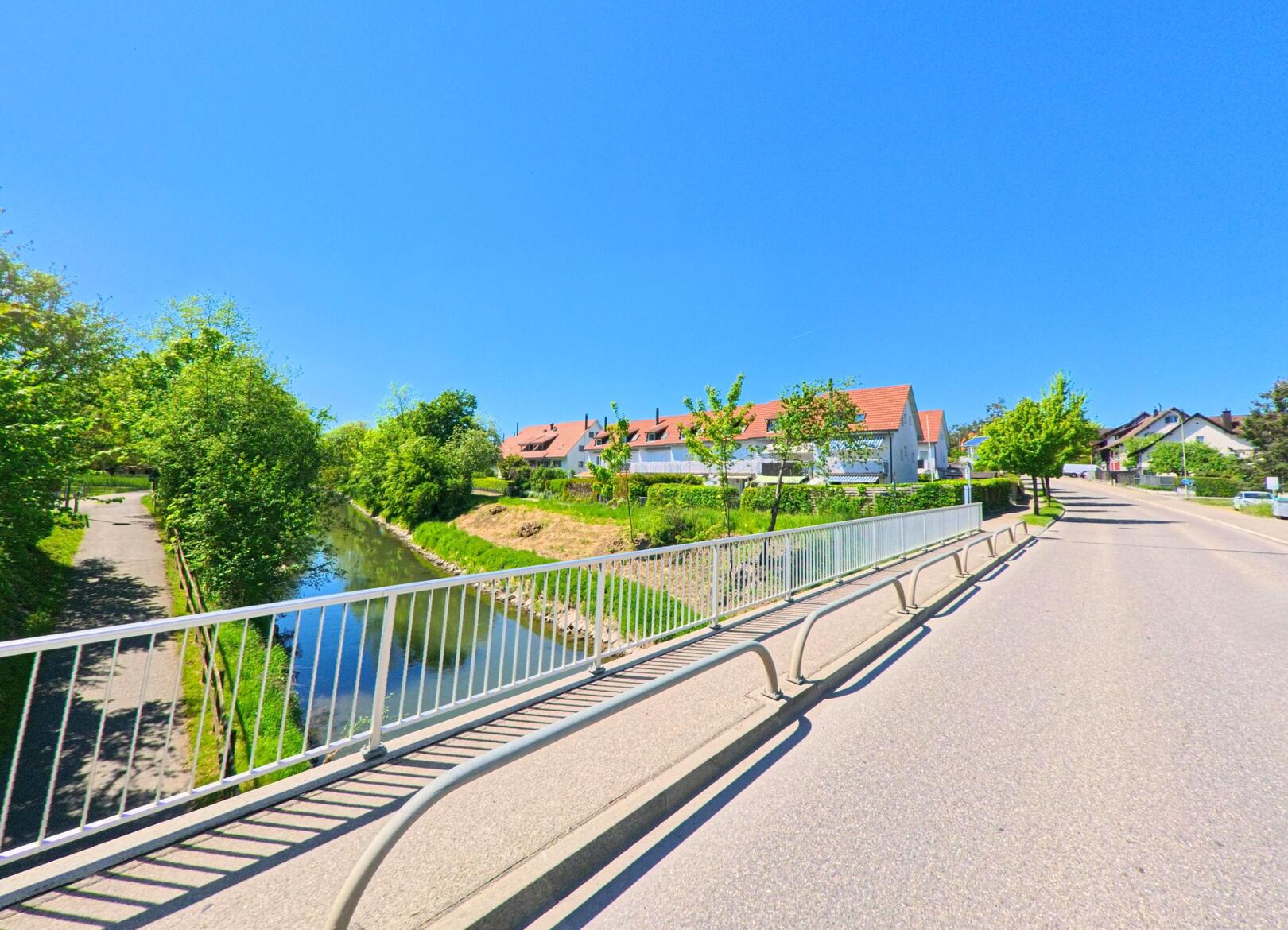 In Oberglatt (8154, Kanton Zürich) fuehrt eine Strasse ueber eine Brucke, die einen kleinen Fluss mit angrenzendem Fussweg ueberquert, waehrend im Hintergrund Wohnhaeuser mit roten Daechern stehen.