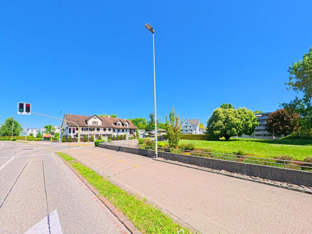 In Oberglatt (8154, Kanton Zürich) steht ein traditionelles Gebaeude mit Balkon an einer Strassenkreuzung mit Ampel, gegenueber einer grossen Gruenflaeche mit Baeumen.