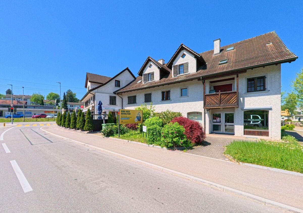 Ein zweistoeckiges Gebaeude mit Balkon und Gastbereich steht in Oberglatt (8154, Kanton Zürich) an einer Strasse nahe einer Kreuzung mit Ampelanlage.