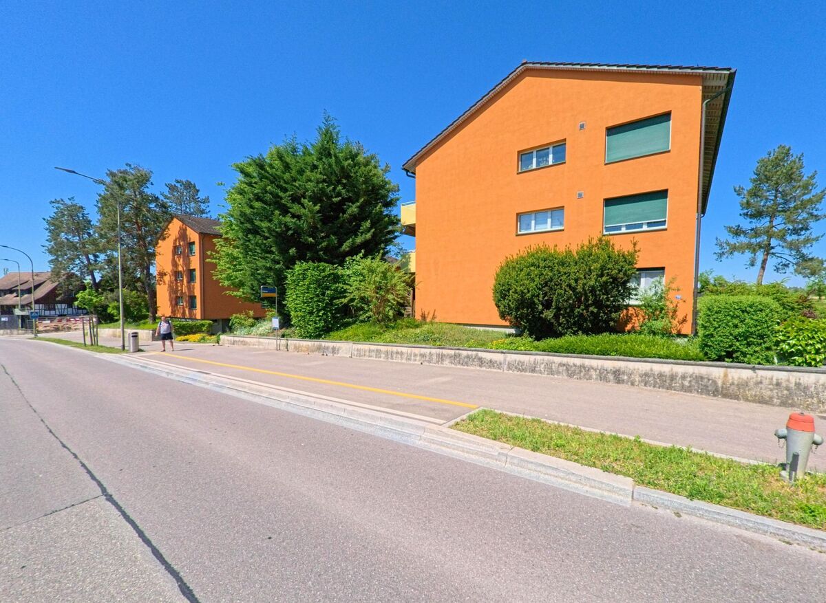 Zwei orange Mehrfamilienhaeuser mit gruener Umrandung stehen in Oberglatt (8154, Kanton Zürich) an einer sonnigen Strasse mit Gehweg.