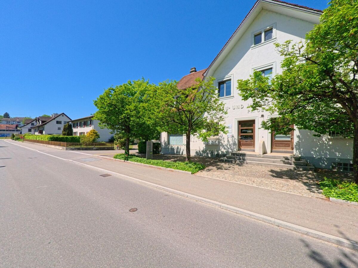 Ein weisses Gebaeude mit der Aufschrift „Primar- und Sekundarschule“ steht in Oberglatt (8154, Kanton Zürich) an einer sonnigen Strasse, gesaeumt von Baeumen.