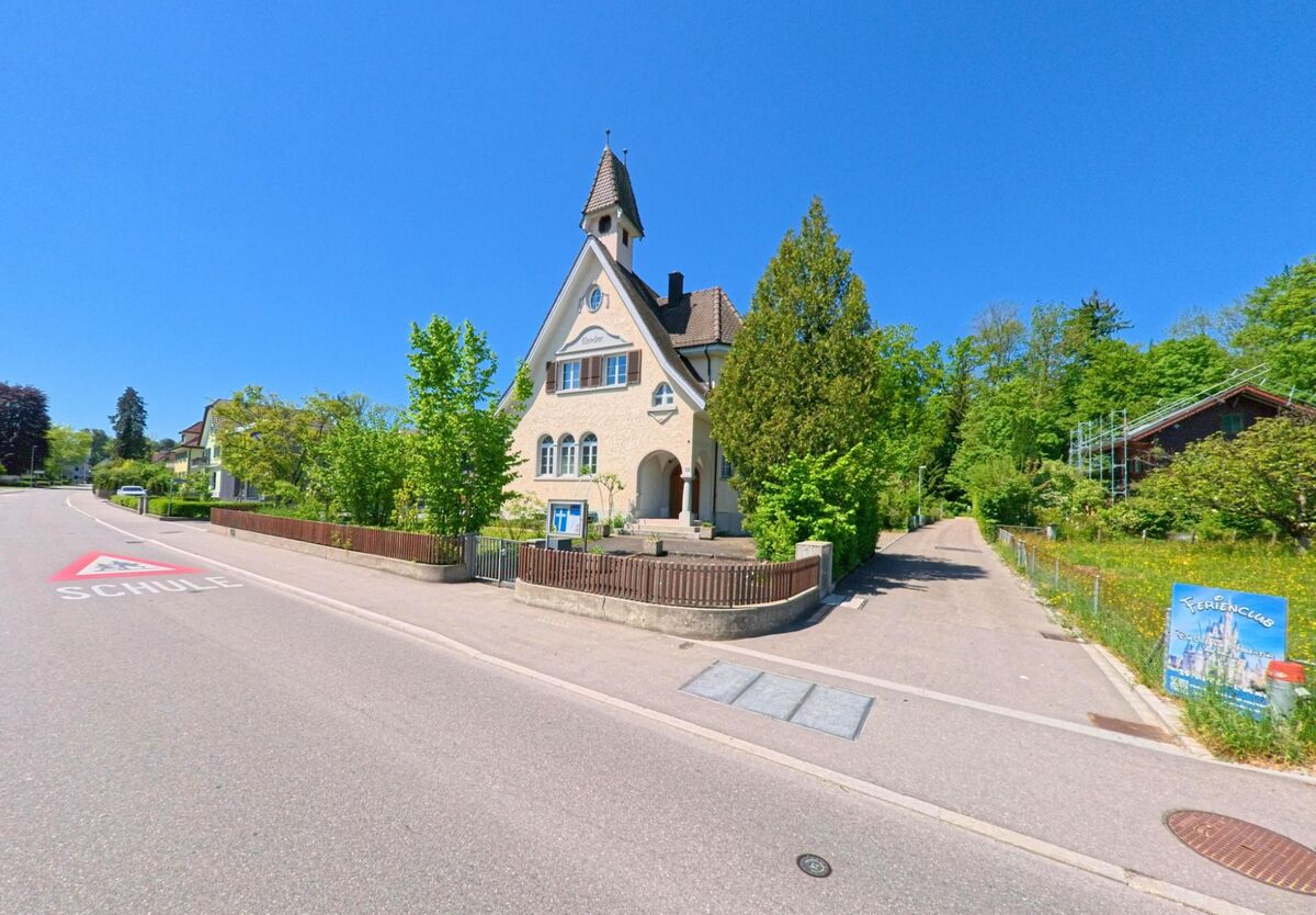 Ein historisches Gebaeude mit Turmspitze dient in Oberglatt (8154, Kanton Zürich) als Schule, gelegen an einer Strasse mit gut sichtbarer Fahrbahnmarkierung.