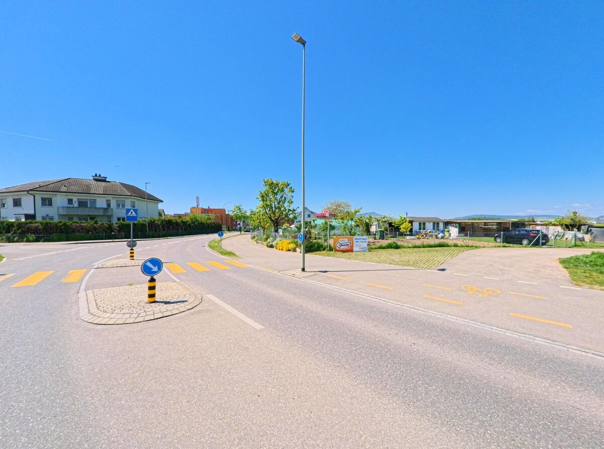 Ein Verkehrsknotenpunkt in Oberglatt (8154, Kanton Zürich) mit Fussgaengerstreifen, Verkehrsinsel und angrenzenden Wohn- und Gartenanlagen unter klarem Himmel.