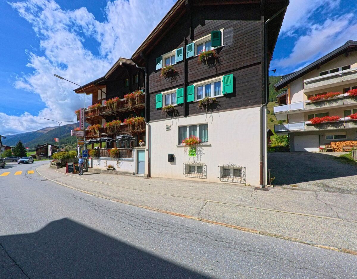 In Obergoms (3988, Kanton Wallis) stehen traditionelle Holzhäuser mit Blumenschmuck entlang einer Strasse unter blauem Himmel.