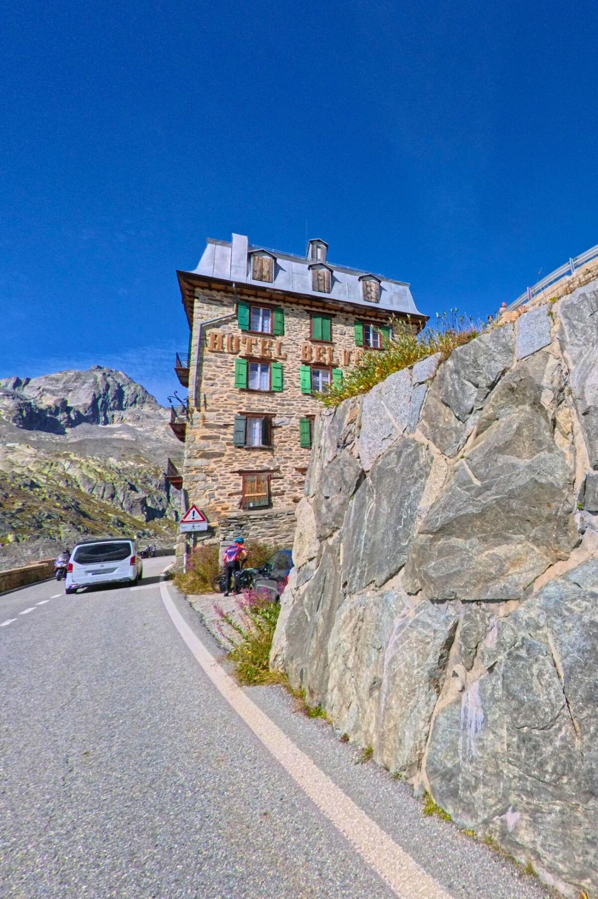 Das Hotel Belvedere in Obergoms (3999, Kanton Wallis) steht direkt an einer Passstrasse vor felsiger Berglandschaft unter klarem Himmel.