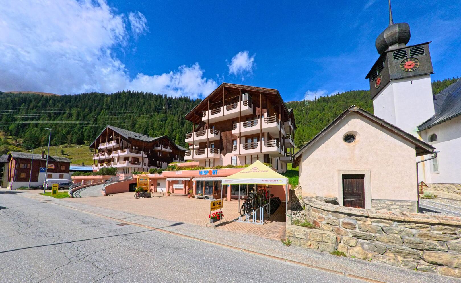 In Obergoms (3999, Kanton Wallis) stehen moderne Wohnhaeuser mit Balkonen neben einer Kirche mit markantem Turm, davor befindet sich ein Fahrradverleih.