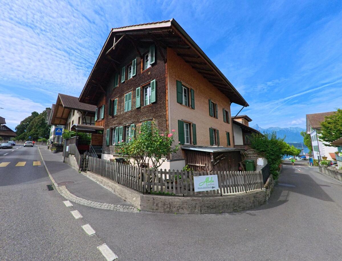 In Oberhofen am Thunersee (3653, Kanton Bern) treffen eine Hauptstrasse mit Zebrastreifen und eine kleine Seitenstrasse bei traditionellen Holzhäusern aufeinander.