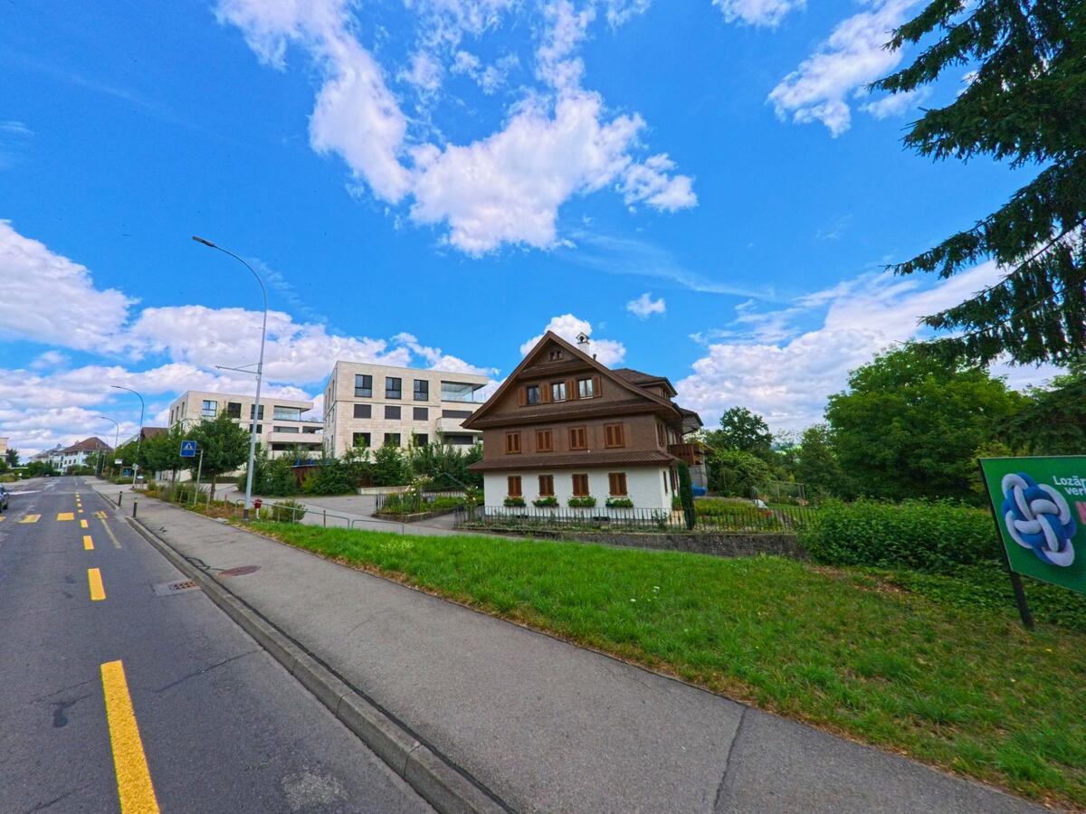In Oberkirch (6208, Kanton Luzern) trifft ein traditionelles Wohnhaus mit Holzfassade auf moderne Mehrfamilienhaeuser entlang einer verkehrsberuhigten Strasse.