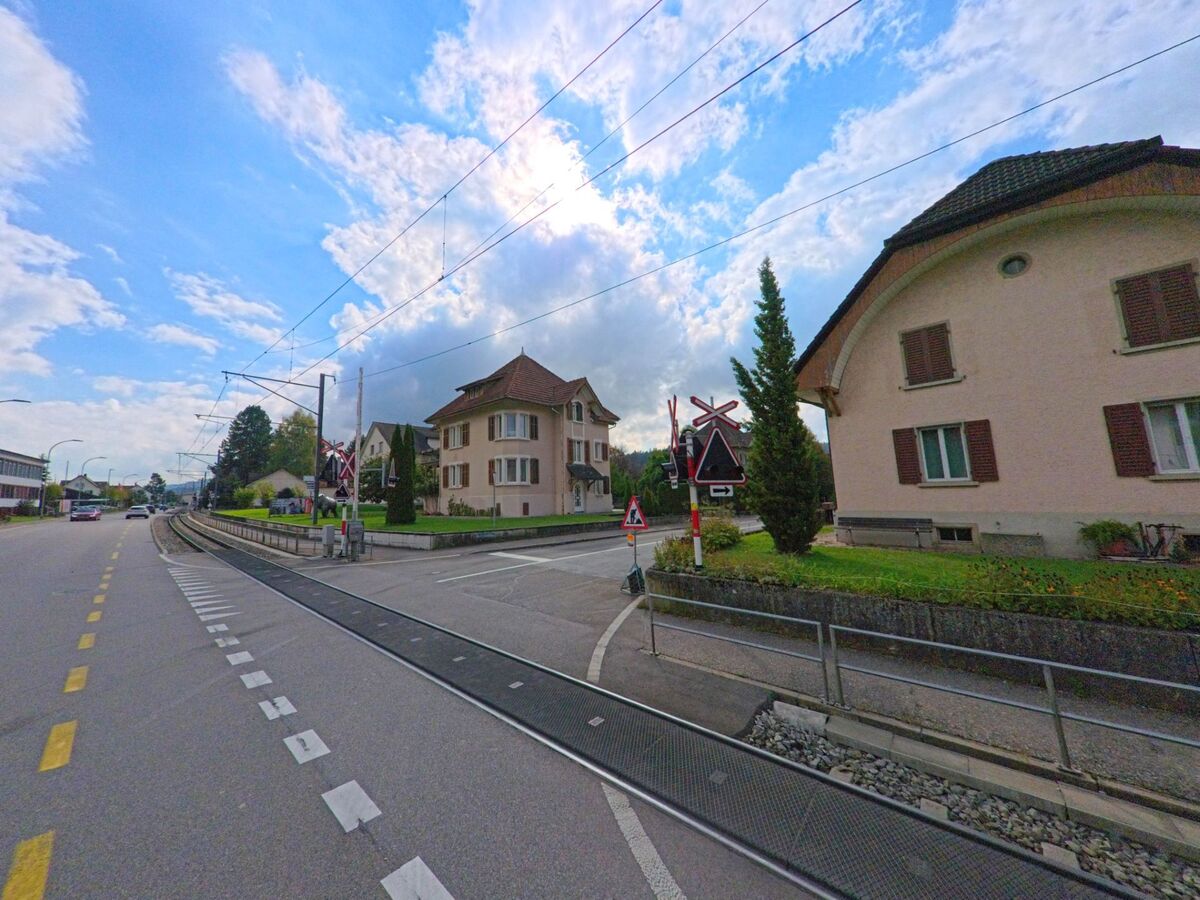 Ein Bahnuebergang mit Schranken fuehrt in Oberkulm (5727, Kanton Aargau) zwischen Wohnhaeusern ueber die Gleise einer Vorortlinie.