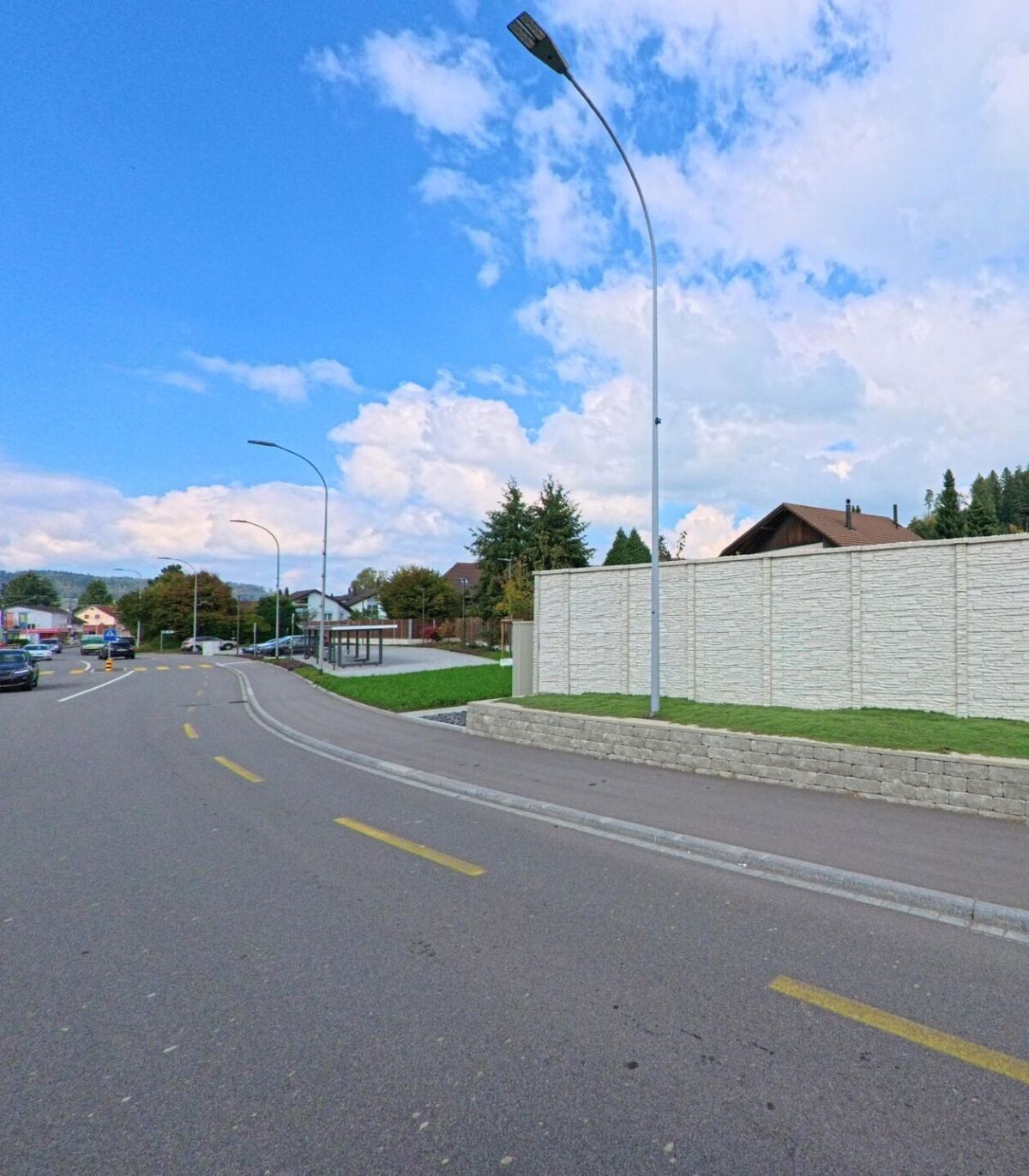 Eine leicht gekruemmte Strasse in Oberkulm (5727, Kanton Aargau) fuehrt an einer Lärmschutzwand und einer Bushaltestelle vorbei unter blauem Himmel.
