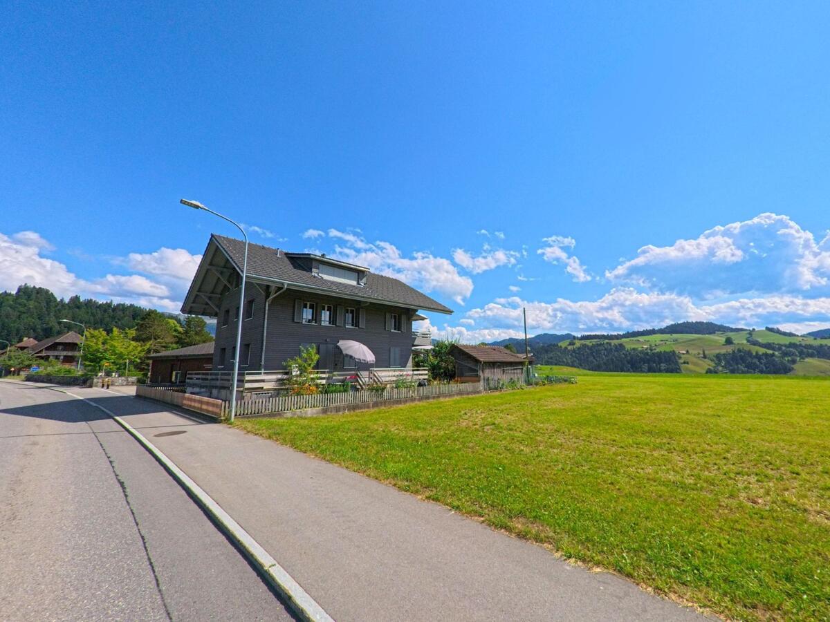 Ein grau gestrichenes Wohnhaus mit umzaeuntem Garten steht in Oberlangenegg (3616, Kanton Bern) am Rand einer ruhigen Strasse mit weitem Blick auf gruene Huegel.
