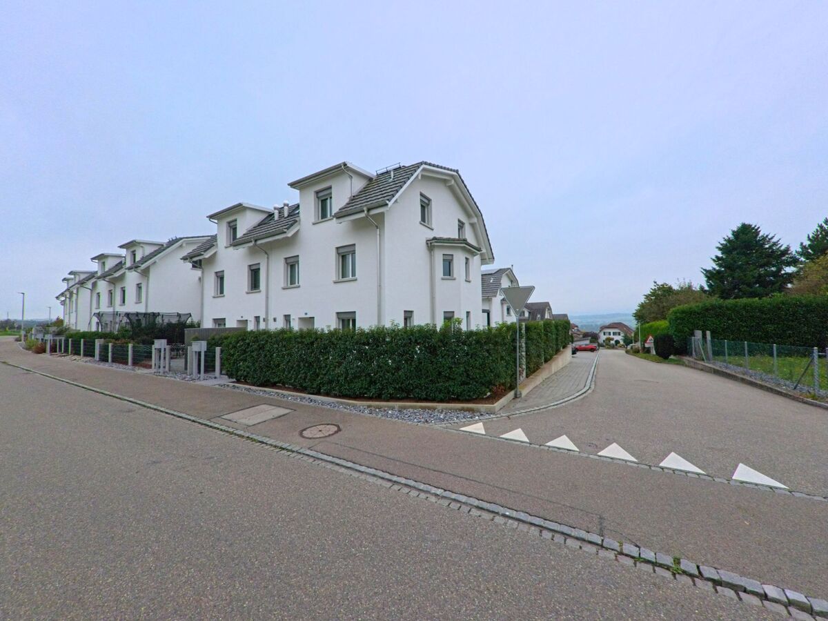 Eine Reihe weisser Doppelhaeuser steht in einer ruhigen Wohnsiedlung von Oberlunkhofen (8917, Kanton Aargau) an einer abfallenden Strasse mit Hecken und Baeumen.