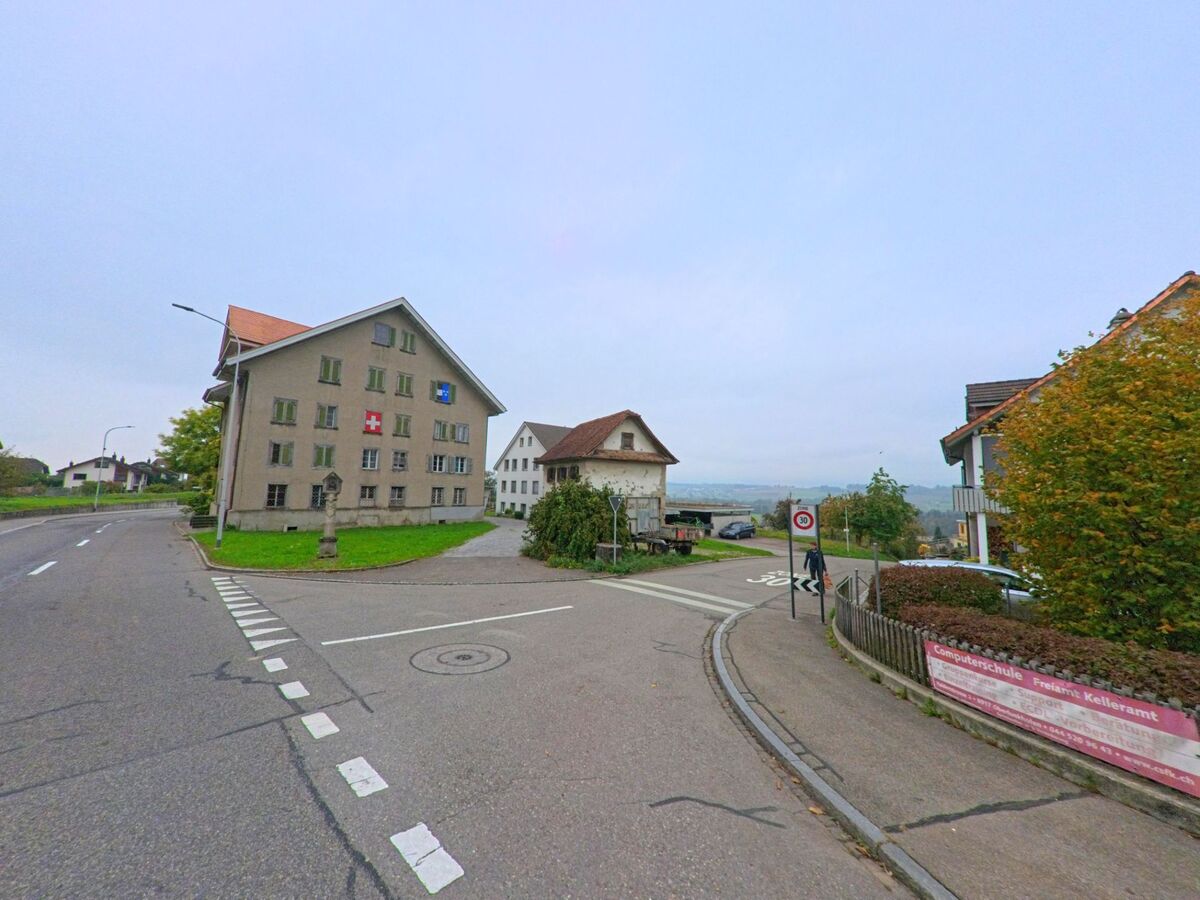 Ein grosses Wohnhaus mit Schweizer und Aargauer Flagge steht an einer Strassenkreuzung in Oberlunkhofen (8917, Kanton Aargau) mit Blick auf das Tal im Hintergrund.