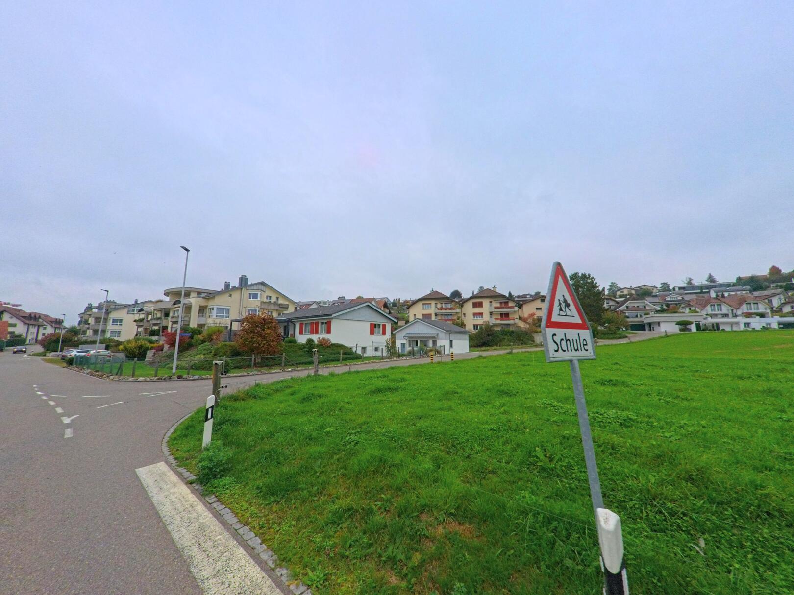 Ein Strassenschild mit dem Hinweis auf eine Schule steht auf einer Gruenflaeche in Oberlunkhofen (8917, Kanton Aargau) vor einer Reihe moderner Wohnhaeuser.