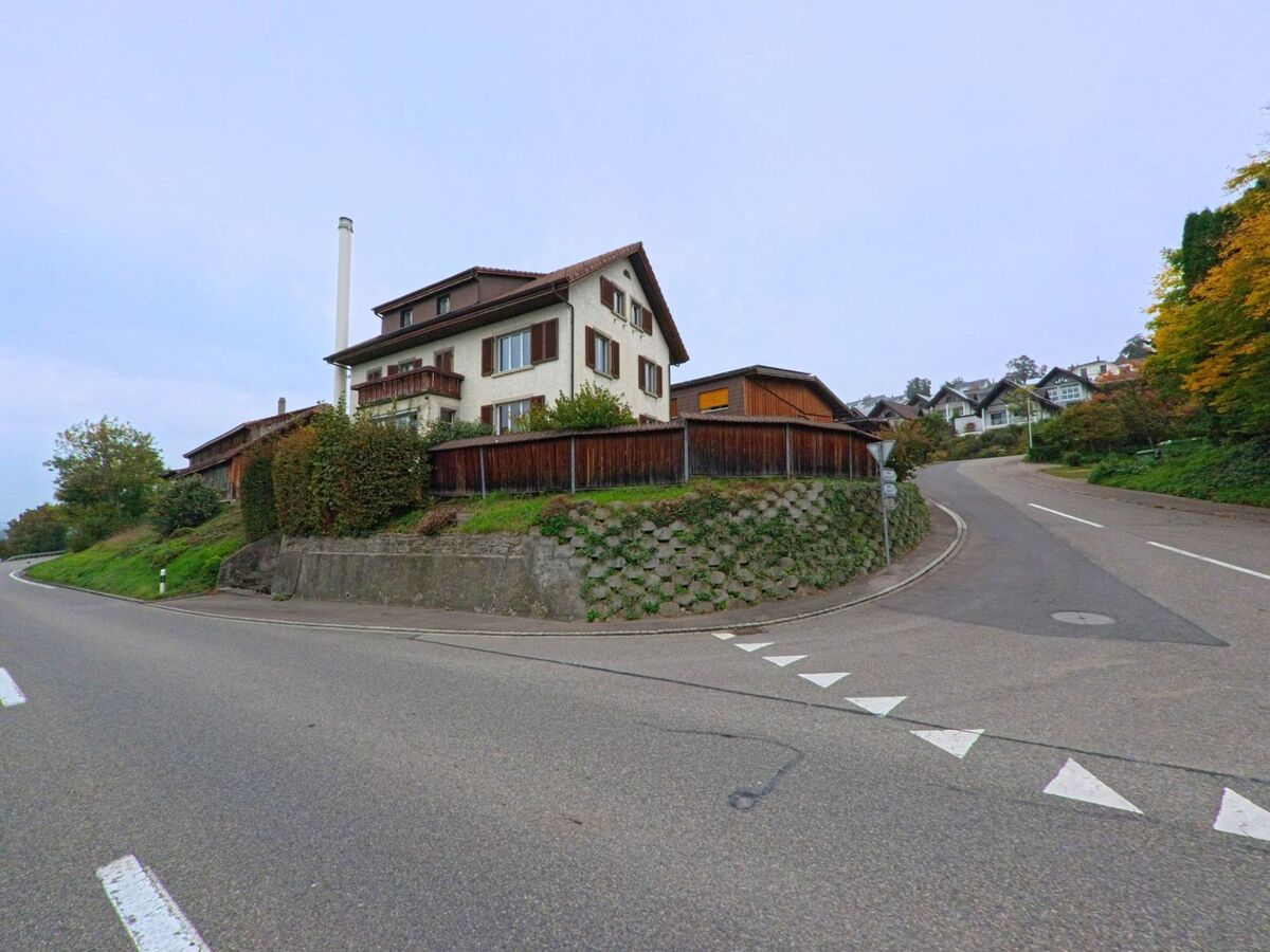 Ein Haus mit braunen Fensterlaeden steht an einer abzweigenden Strasse in Oberlunkhofen (8917, Kanton Aargau), umgeben von weiteren Wohnhaeusern am Hang.