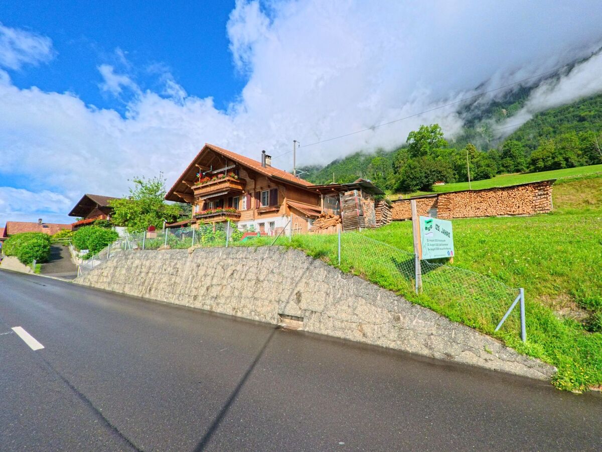 In Oberried am Brienzersee (3854, Kanton Bern) steht ein traditionelles Bauernhaus mit Holzvorräten und Gartenschild am Rand einer nassen Landstrasse vor bewaldetem Hang.