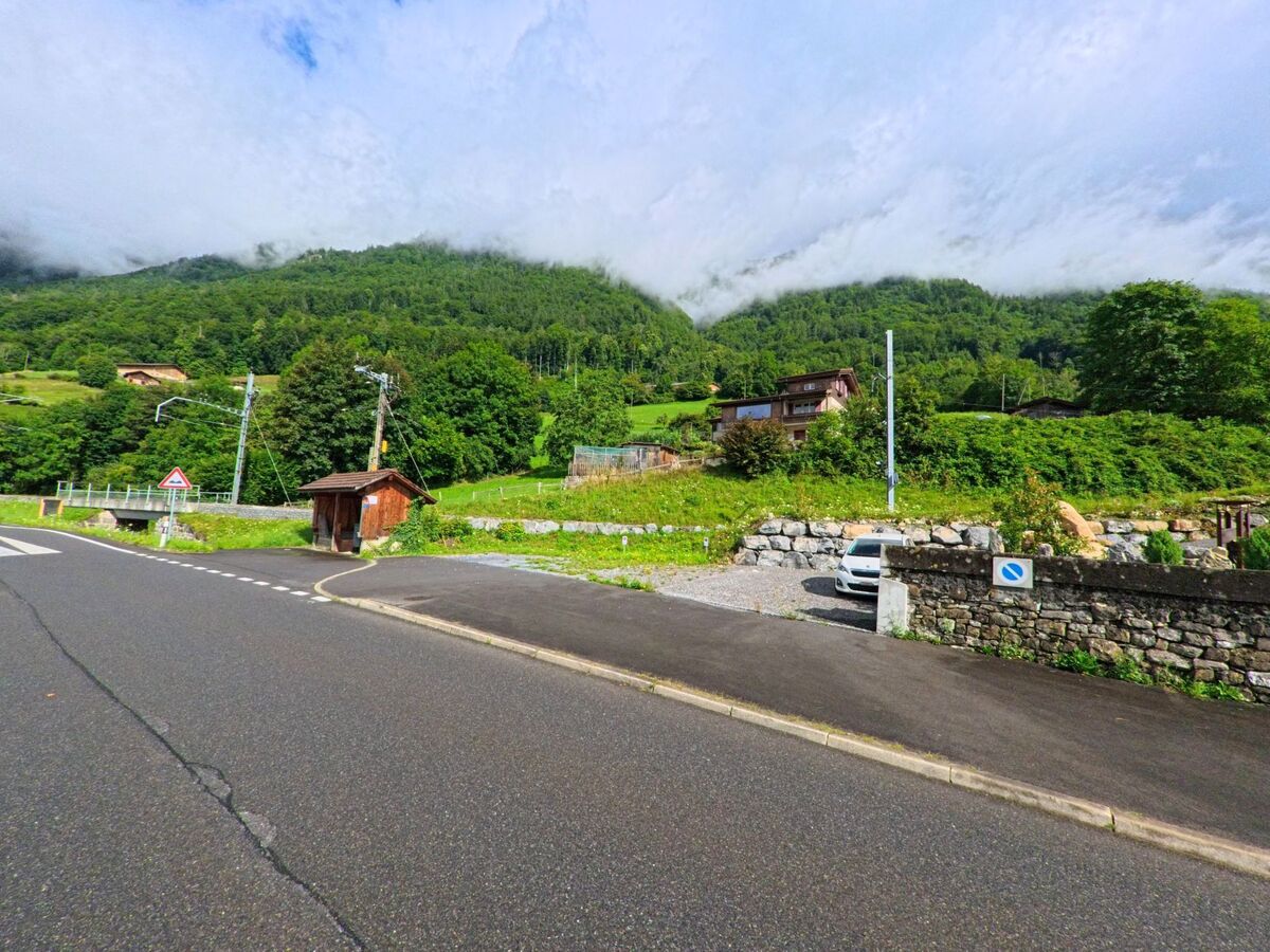 In Oberried am Brienzersee (3854, Kanton Bern) trifft eine Seitenstrasse auf die Hauptstrasse vor einem kleinen Holzunterstand, umgeben von Wiesen und bewaldeten Hügeln.
