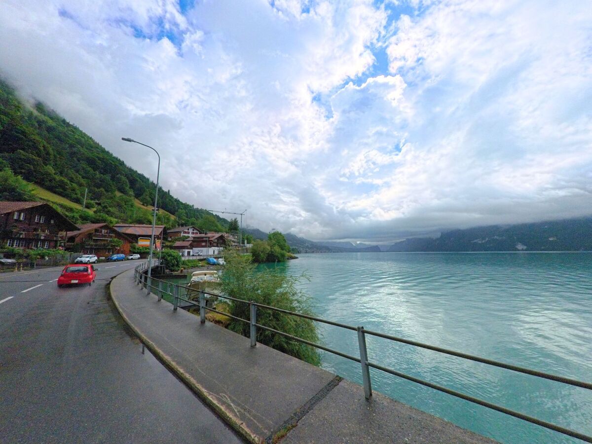 In Oberried am Brienzersee (3854, Kanton Bern) verläuft die Uferstrasse direkt am Brienzersee entlang und führt durch das Dorf mit Blick auf bewaldete Hänge und tiefe Wolken.