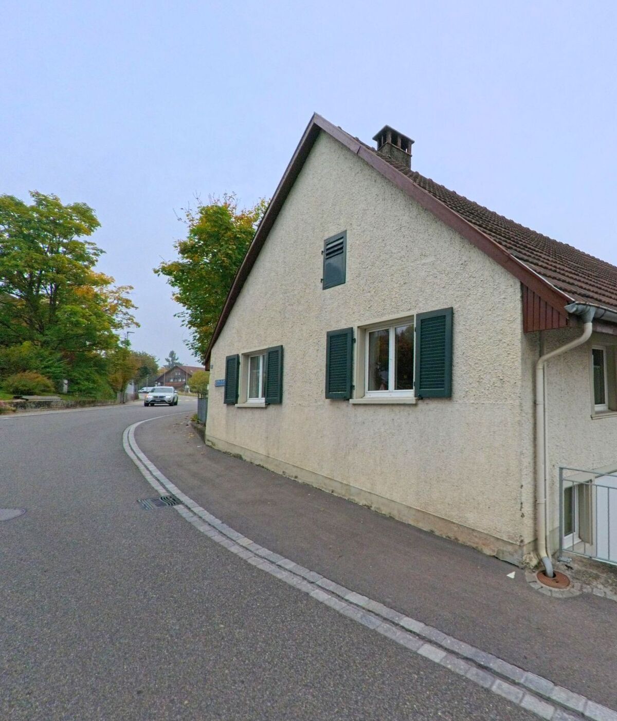 Ein kleines Wohnhaus mit grünen Fensterlaeden steht an einer leicht gekruemmten Strasse in Oberrohrdorf (5452, Kanton Aargau) neben herbstlichen Baeumen.