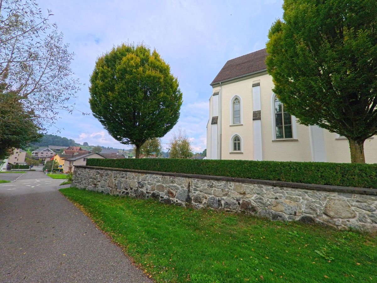 Ein Fussweg in Oberrueti (5647, Kanton Aargau) fuehrt an der Kirche mit ihrer Steinmauer und gepflegten Baeumen vorbei.