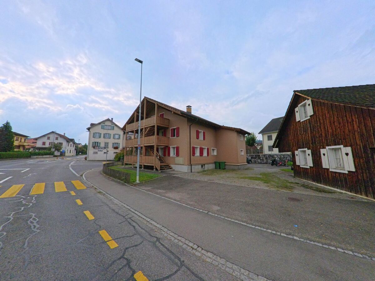 In Oberrueti (5647, Kanton Aargau) stehen traditionelle Wohnhaeuser mit roten Fensterlaeden entlang einer Kreuzung mit Fussgaengerstreifen.