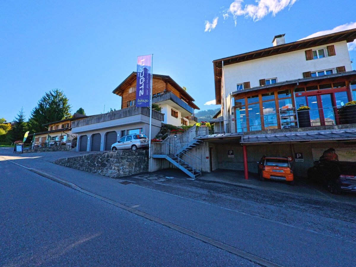 An einer leicht ansteigenden Strasse in Obersaxen-Mundaun (7134, Kanton Graubuenden) stehen mehrere Gebäude mit Garagen und Balkonen, darunter ein Haus mit grosser Glasfront.