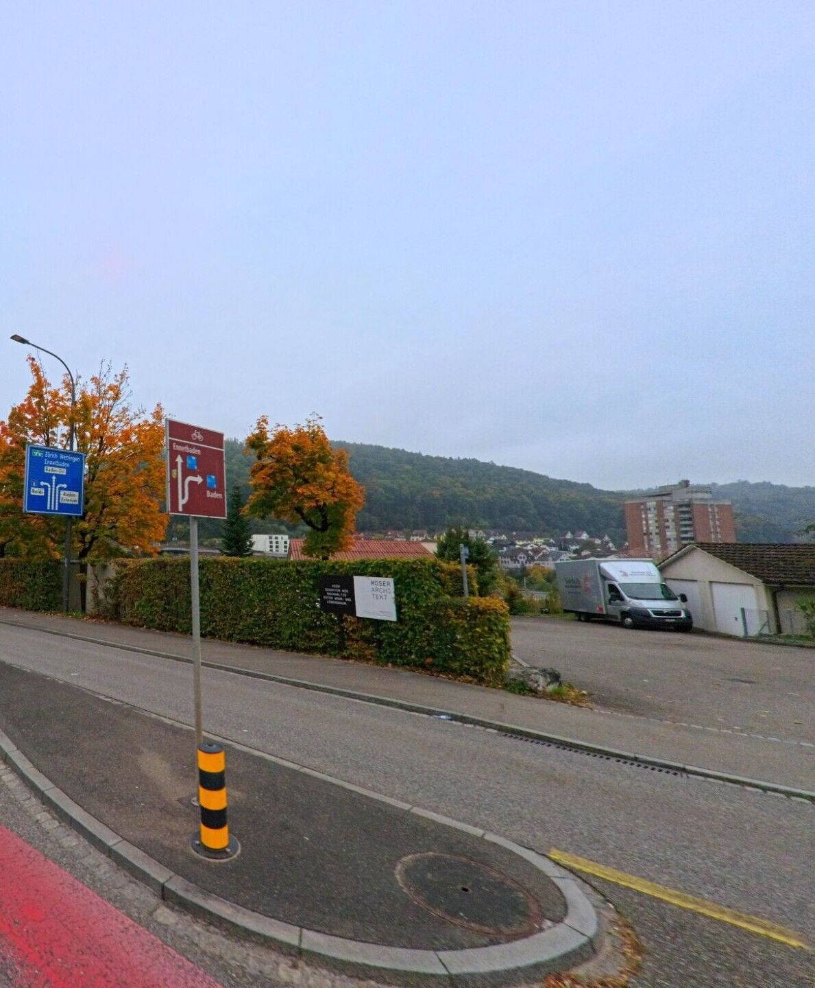 Von einer Strasse in Obersiggenthal (5415, Kanton Aargau) aus sieht man Wegweiser Richtung Baden und Ennetbaden, flankiert von herbstlich gefaerbten Baeumen.