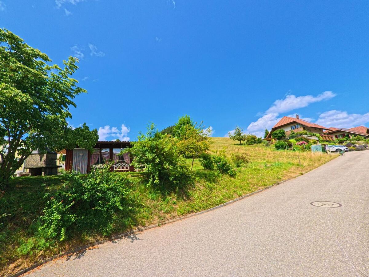 Entlang einer ansteigenden Strasse in Oberthal (3531, Kanton Bern) liegen ein Gartenhaus mit Sitzbank im Schatten sowie ein Wohnhaus auf einer sonnigen Wiese.