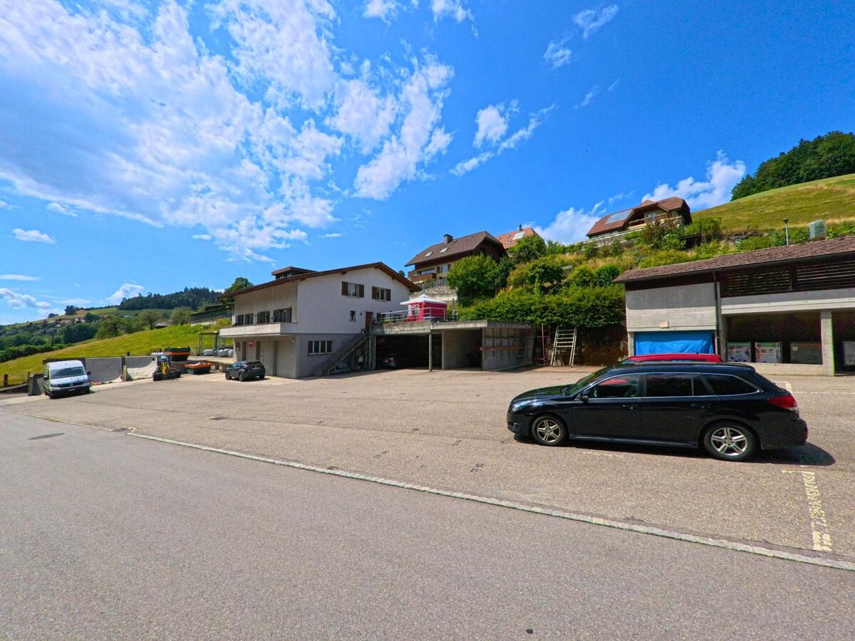 In Oberthal (3531, Kanton Bern) erstreckt sich ein grosser asphaltierter Platz vor einem Wohn- und Gewerbegebäude, umgeben von Hügelhäusern und grüner Landschaft
